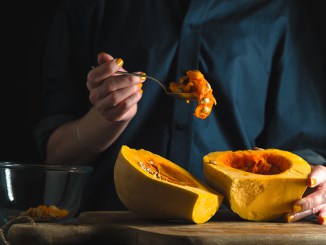 Spoon with seeds in a hand, pumpkin halves on a table on a blue background