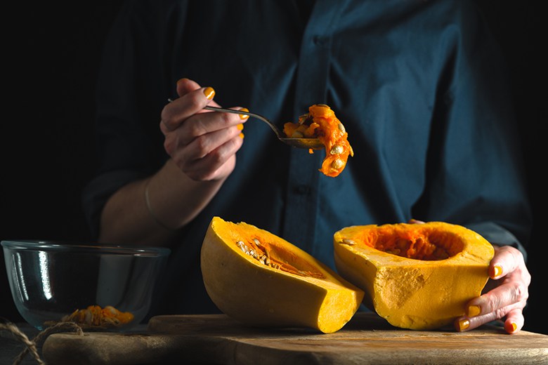 Healthy Kitchen Hacks for the Holidays | Food & Nutrition | November-December 2017 Spoon with seeds in a hand, pumpkin halves on a table on a blue background