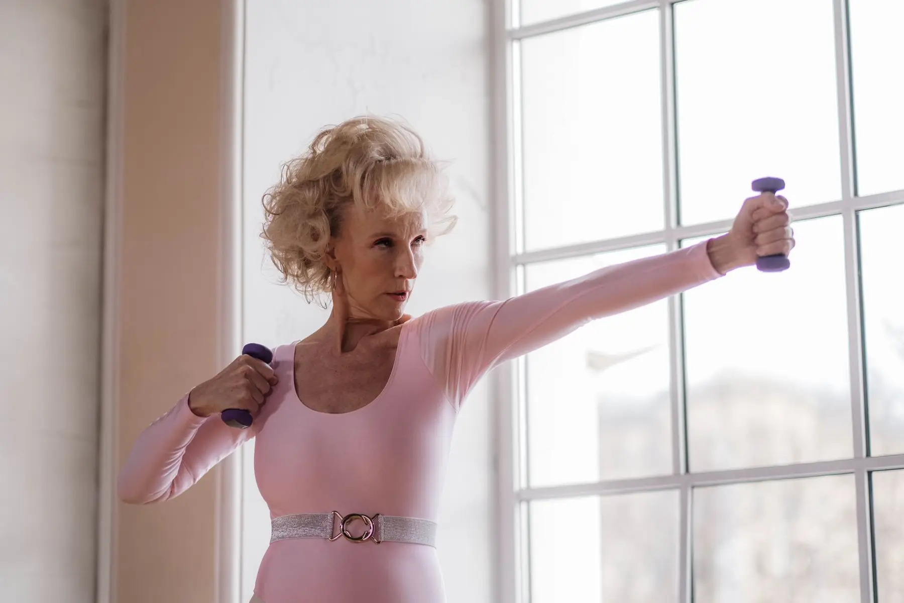 Senior woman in pink leotard working out with dumbbells near a large window indoors